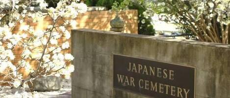 Un cartel que dice 'Japanese War Cemetery' con un muro de piedra y árboles en flor al fondo.