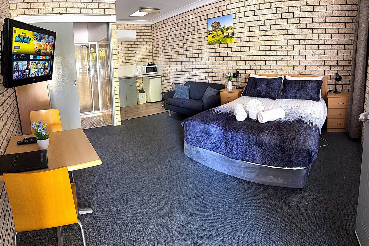 A hotel room with a bed, a desk, a TV, a small kitchenette, and a brick wall.