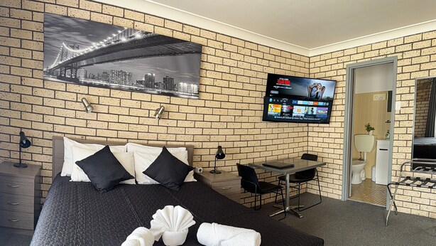 A hotel room with a brick wall, a bed with black and white bedding, a TV mounted on the wall, a desk with a chair, and a bathroom visible through an open door. - Cowra Crest Motel (Cowra)