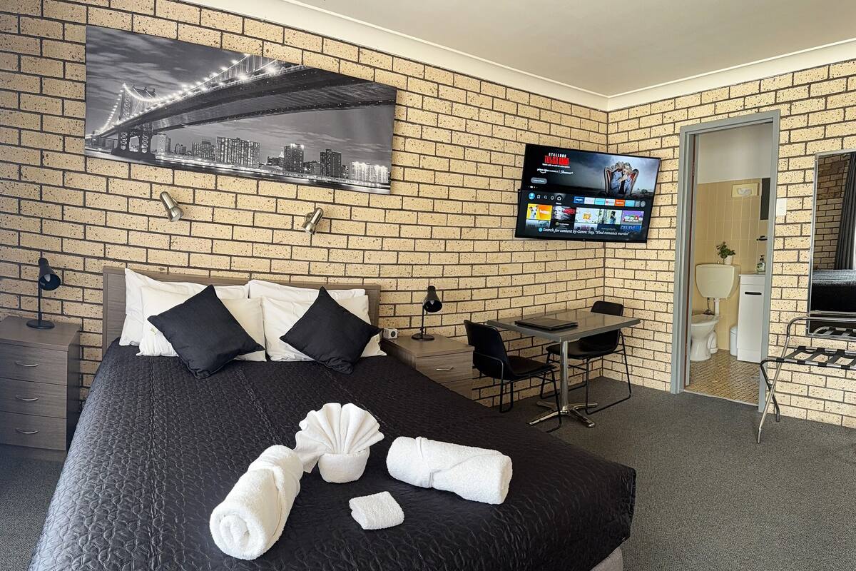 A hotel room with a brick wall, a bed with black and white bedding, a TV mounted on the wall, a desk with a chair, and a bathroom visible through an open door.