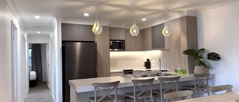 A modern kitchen with a central island, bar stools, a dining table, pendant lighting, and a refrigerator.