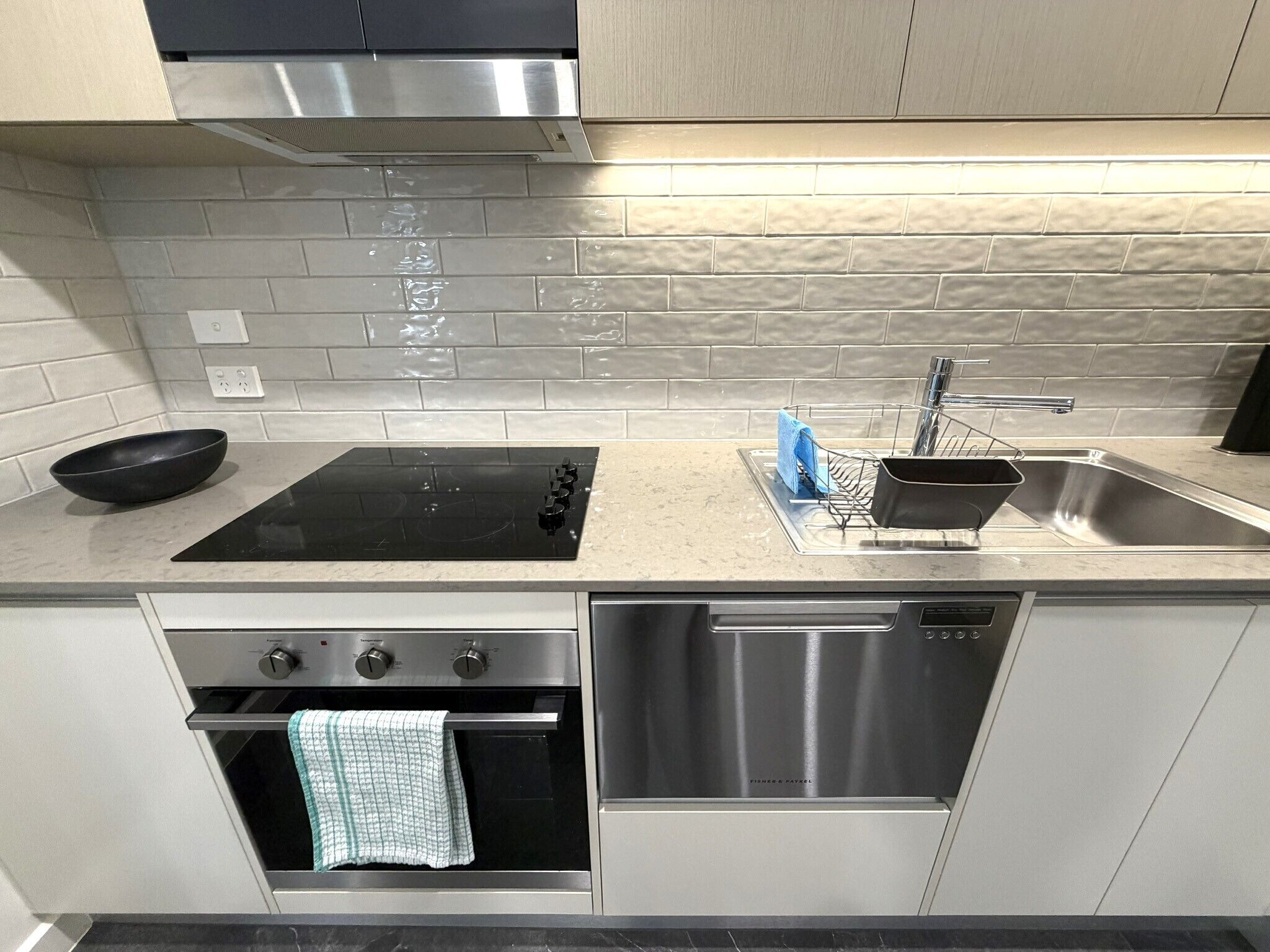 Una cocina moderna con cocina por inducción negra, pileta de acero inoxidable, lavavajillas, horno y salpicadero de azulejos.