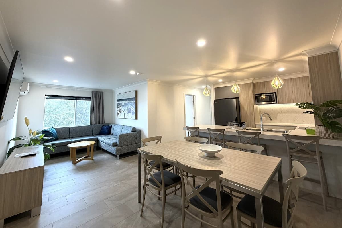 A modern kitchen with a central island, a dining area with a wooden table and chairs, a seating area with a sofa and a small coffee table, and a large window providing a view.