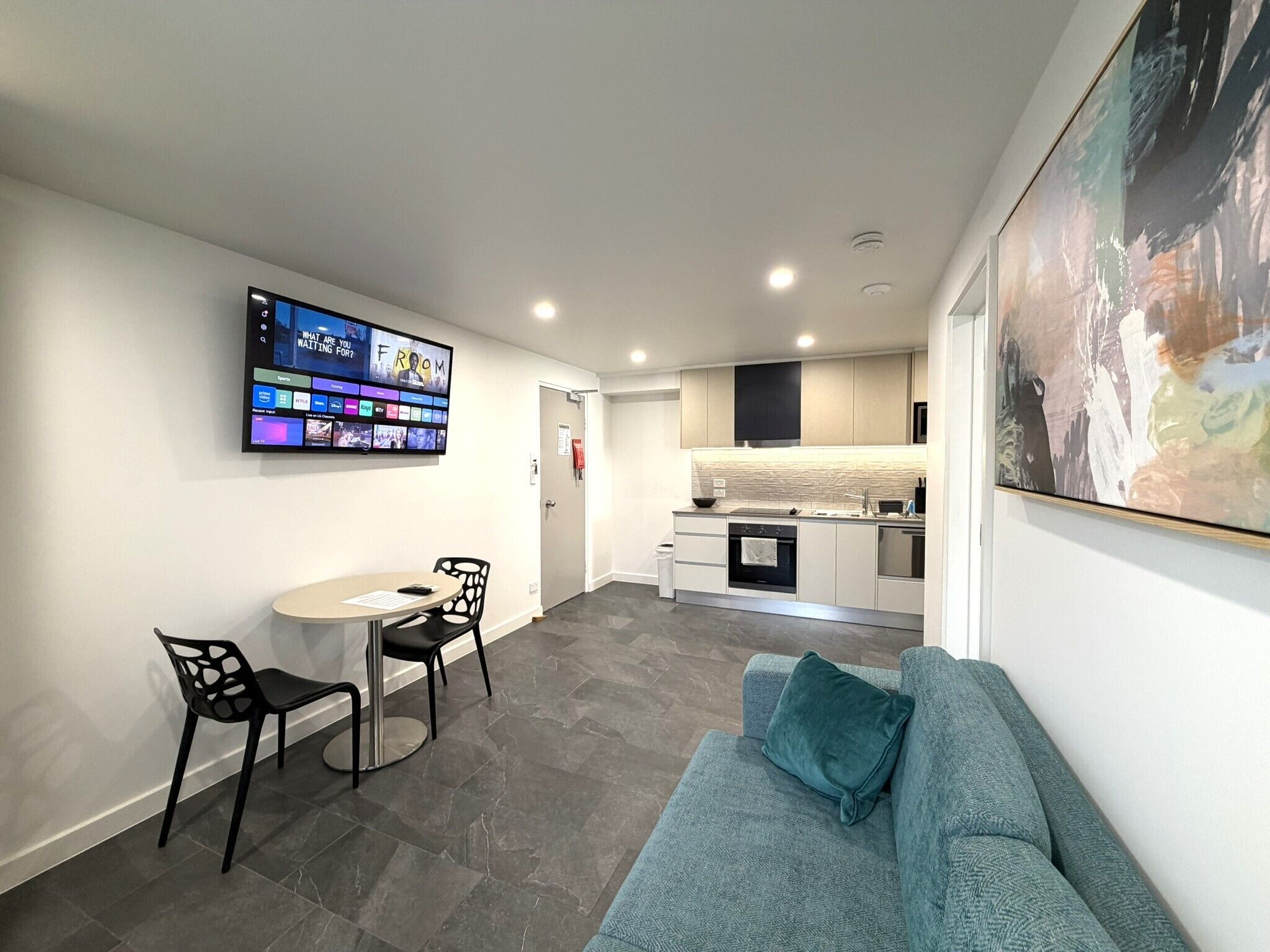 A modern living room with a sofa, a TV mounted on the wall, and a kitchen in the background.
