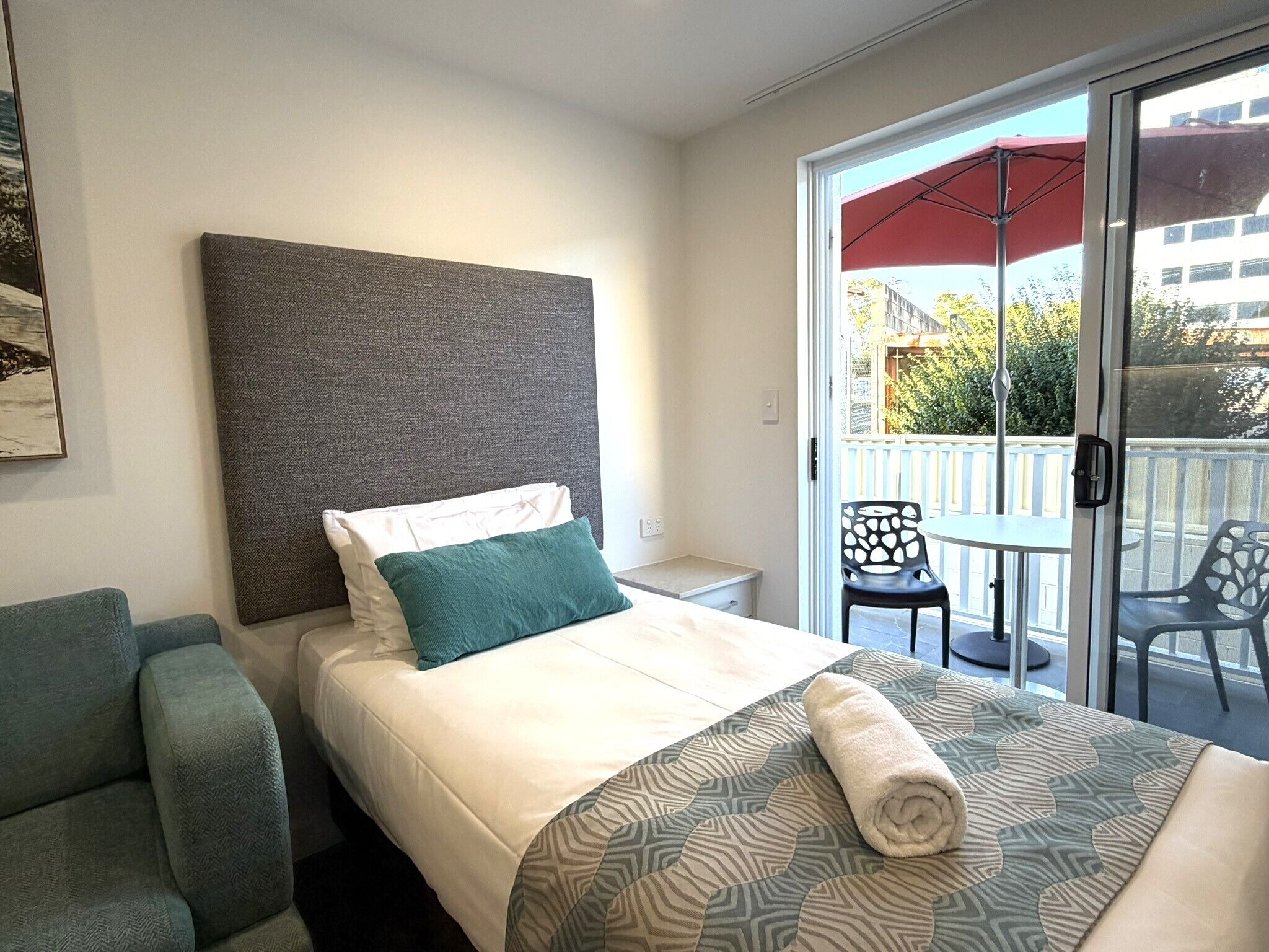 Habitación de hotel con una cama, una silla, una mesita y un balcón con una sombrilla roja.