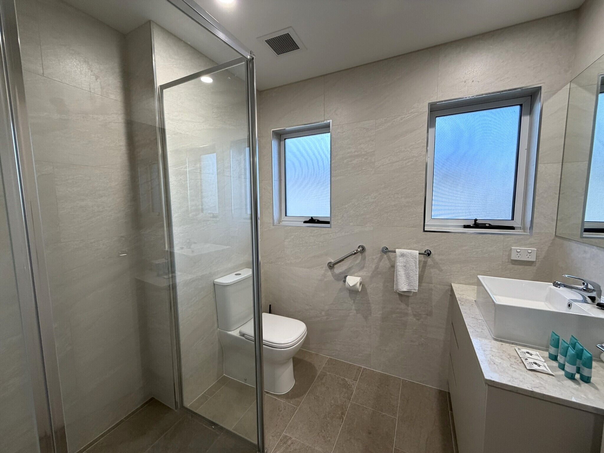 A modern bathroom with a glass shower enclosure, a toilet, a sink with a mirror, and two windows.