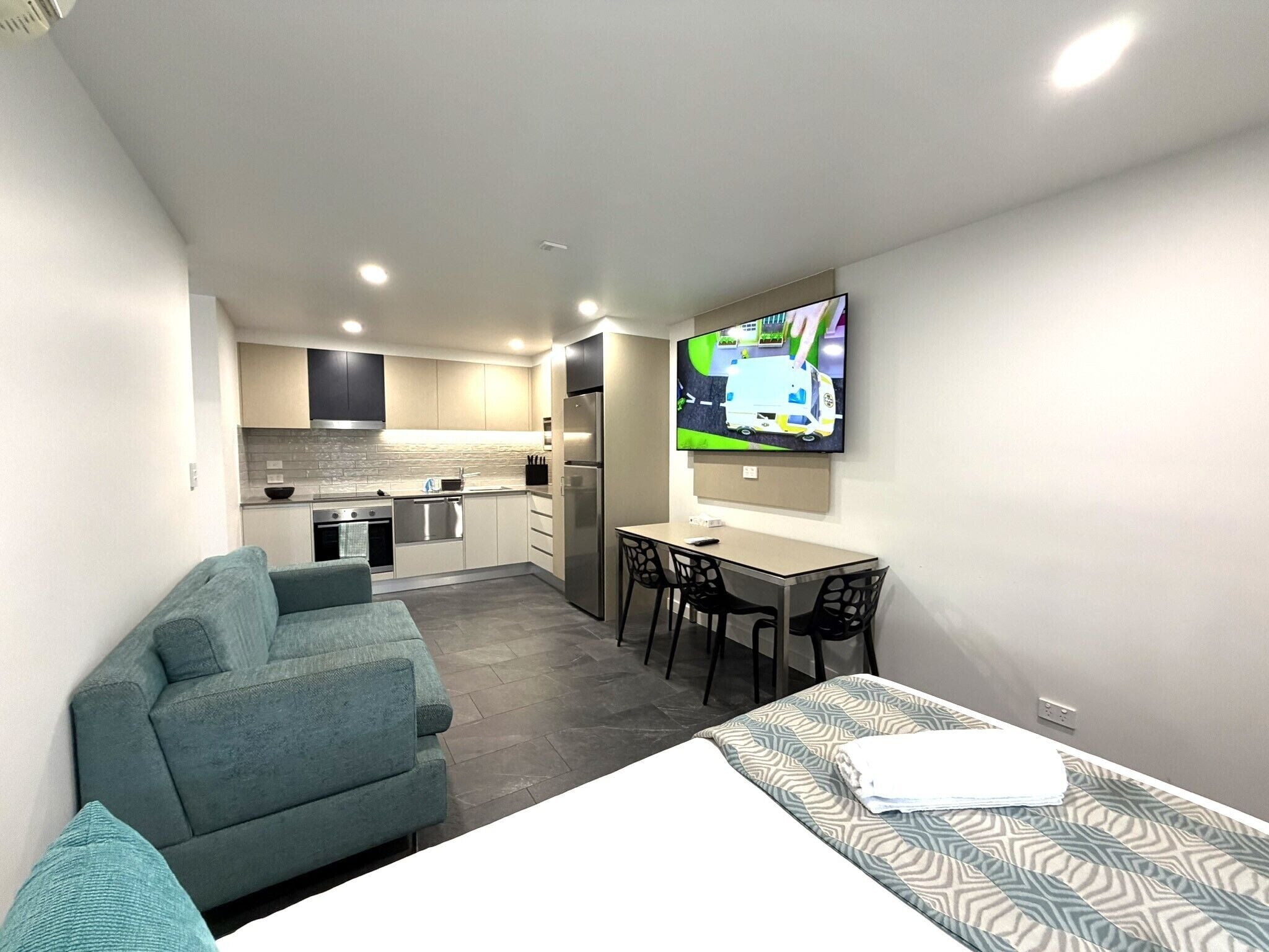 Una habitación de hotel compacta con kitchenette, un sofá, una mesa de comedor con sillas y un televisor montado en la pared.