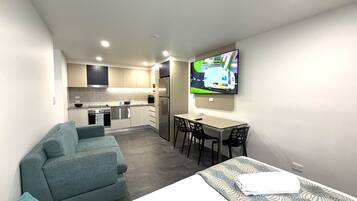 A compact hotel room with a kitchenette, a sofa, a dining table with chairs, and a TV mounted on the wall.
