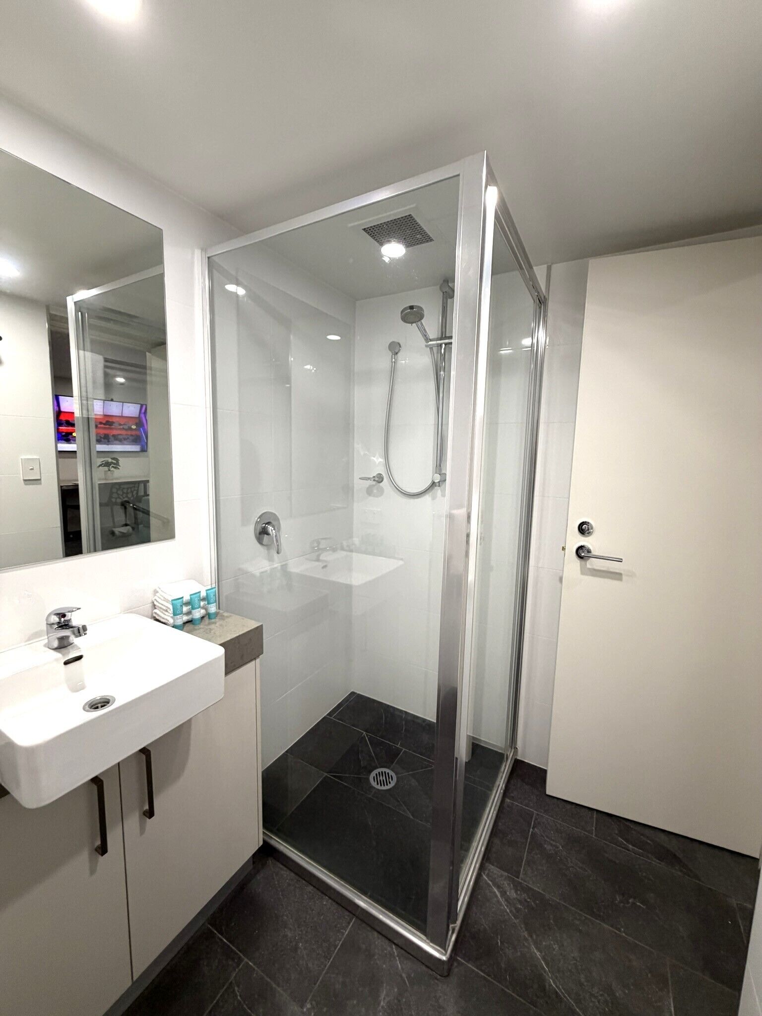 Un baño moderno con ducha envuelta en vidrio, lavamanos con espejo y suelo de baldosas oscuras.