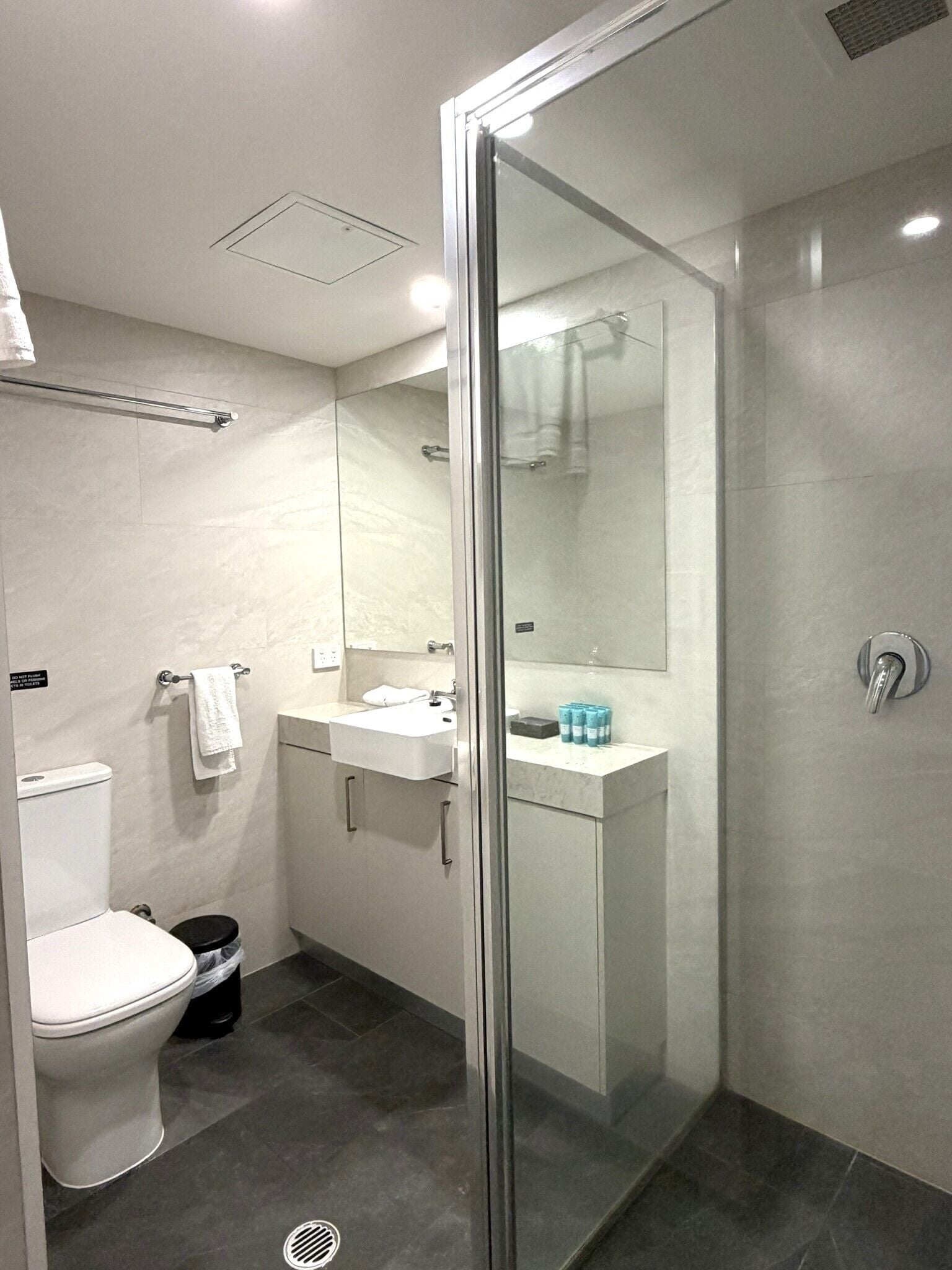 Un baño moderno con ducha de vidrio, inodoro blanco, lavamanos con espejo y un perchero para toallas.