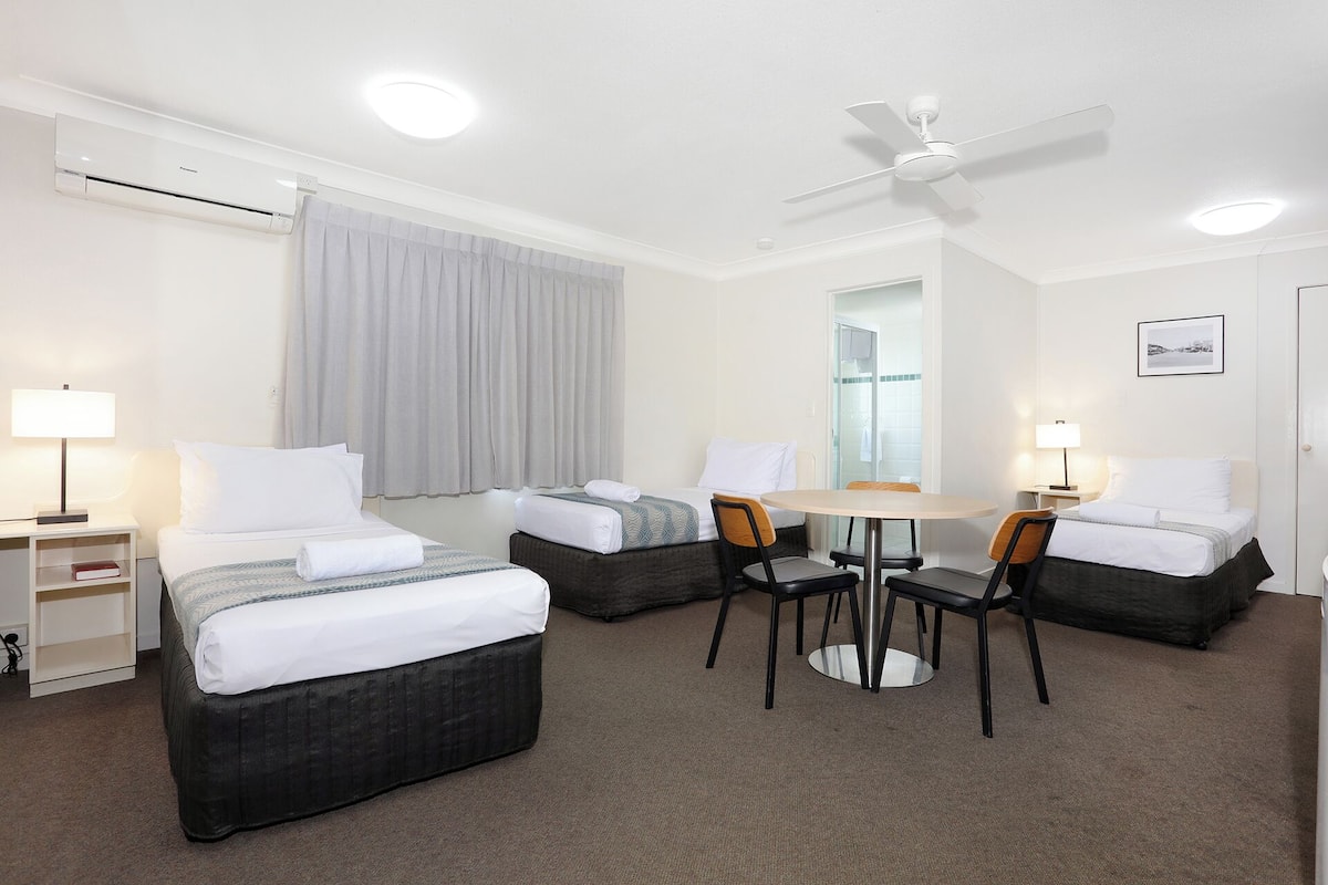 A hotel room with two beds, a dining table, chairs, a ceiling fan, and a window with curtains.