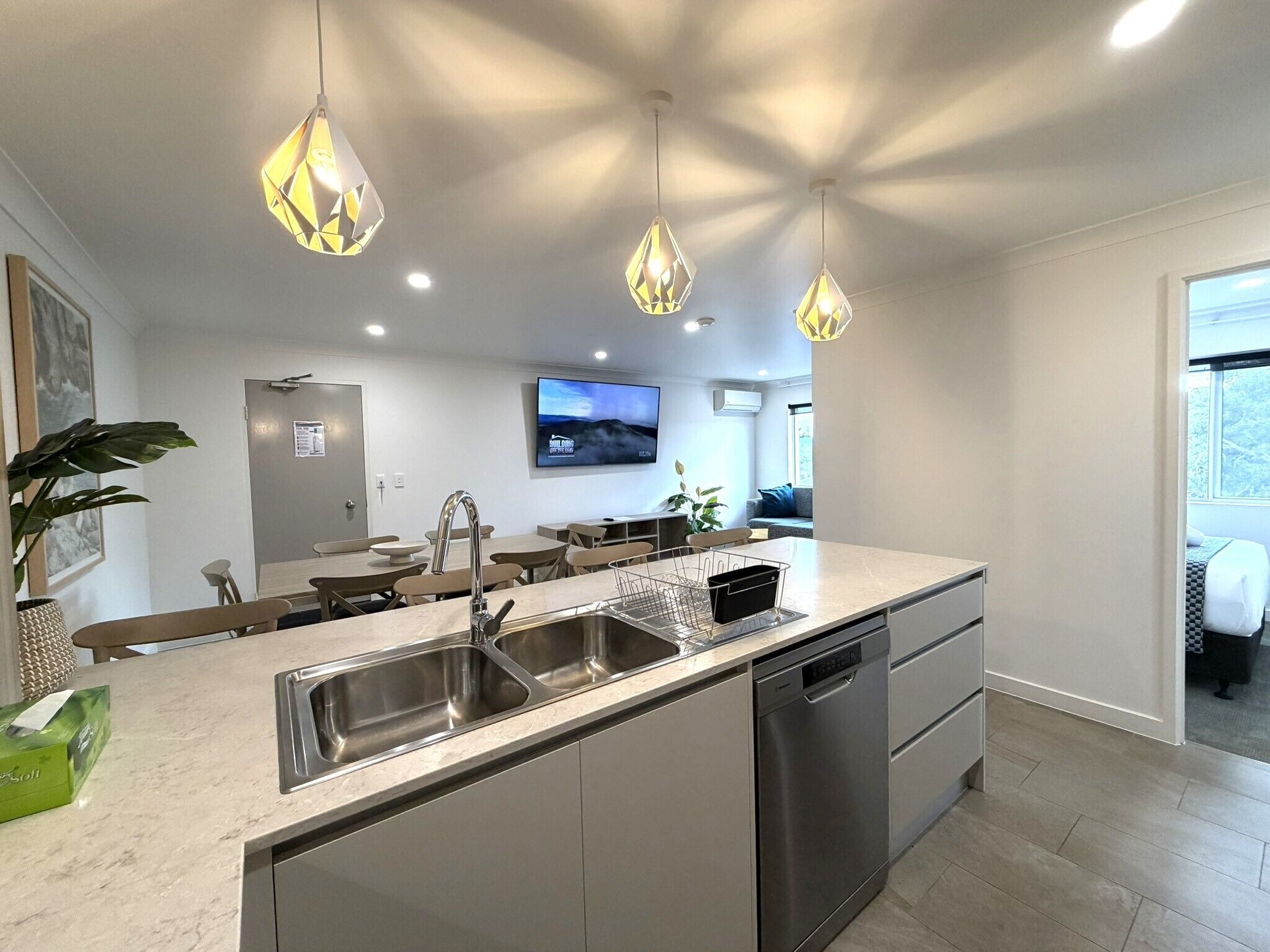 Una cocina moderna con una isla central, electrodomésticos de acero inoxidable, iluminación colgante y un televisor de pantalla plana montado en la pared.