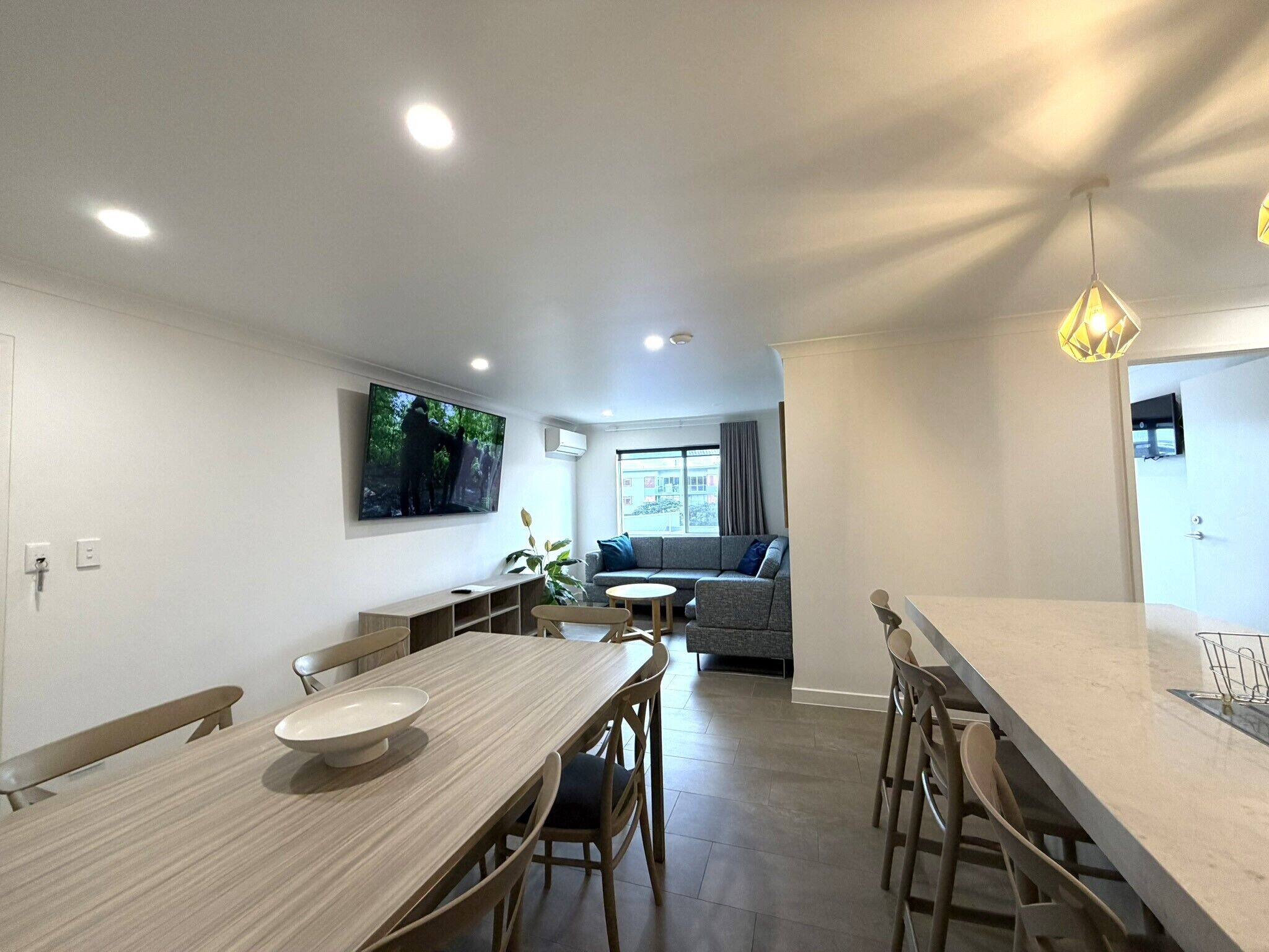 A modern kitchen with a central island, a dining area with a wooden table and chairs, a living room with a sofa and a TV mounted on the wall.