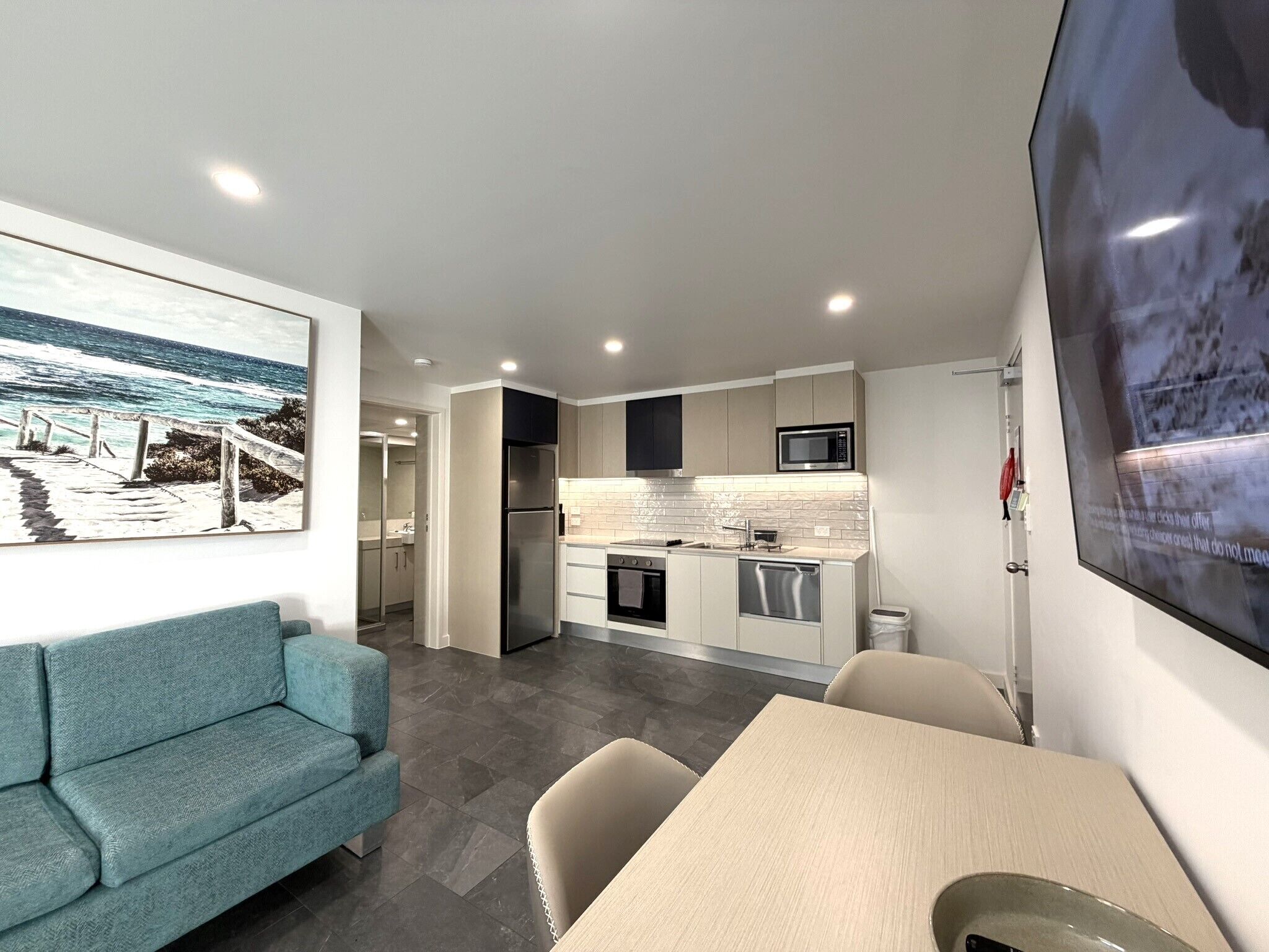 Una cocina moderna con microondas, horno y lavavajillas, un comedor con mesa y sillas, y una sala de estar con sofá y un cuadro de una playa.