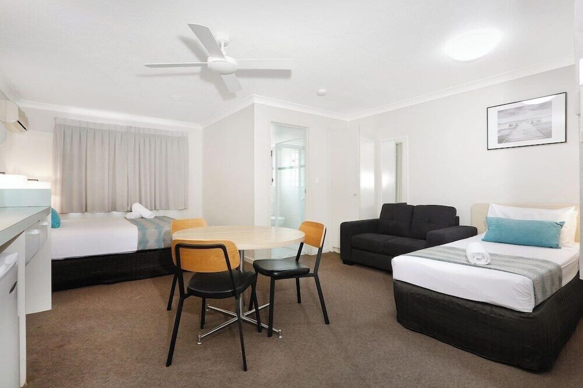 A hotel room with a bed, a sofa, a dining table with chairs, a ceiling fan, and a window.