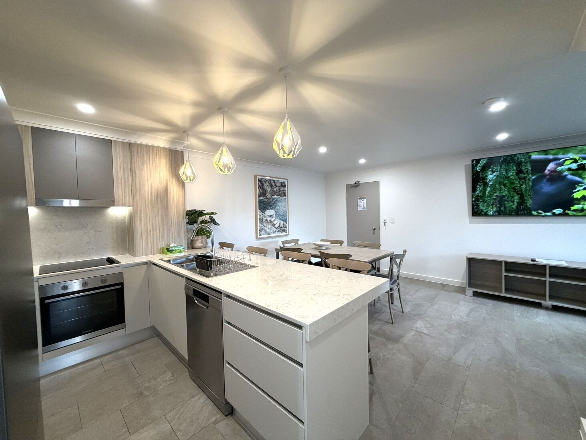Una cocina moderna con una isla central, luces colgantes, una zona de comedor con sillas y un televisor de pantalla plana montado en la pared.