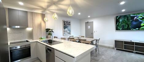 Une cuisine moderne dotée d’un îlot central, de luminaires suspendus, d’un coin repas avec des chaises et d’un téléviseur à écran plat fixé au mur.