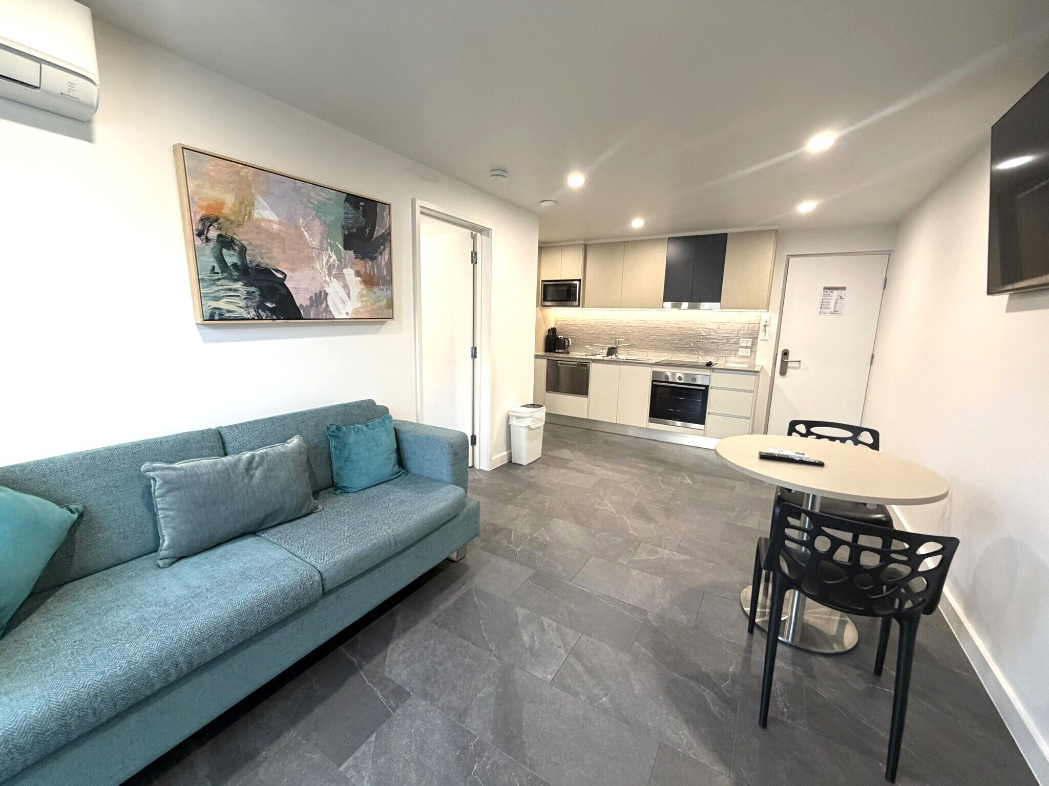 Una sala de estar moderna con un sofá, una mesa de comedor con sillas, una zona de cocina con electrodomésticos y un cuadro en la pared.