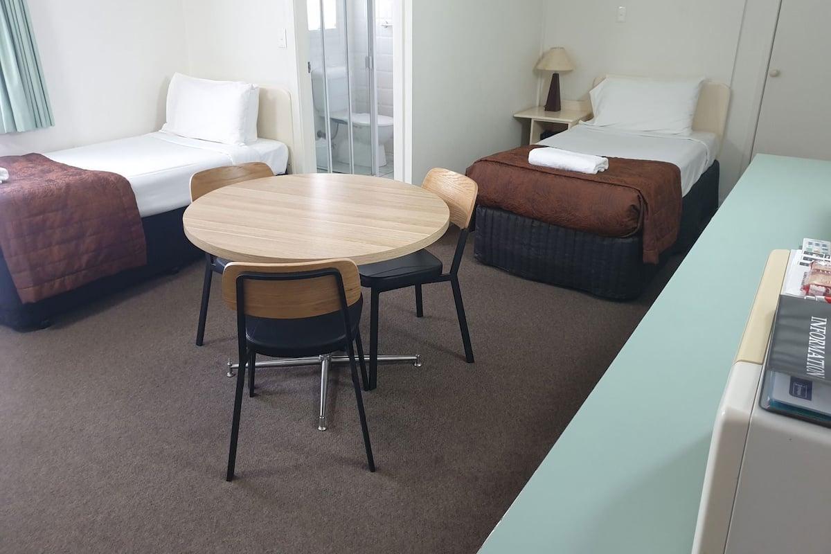 A hotel room with two single beds, a round wooden table with chairs, a small desk, and a minibar.