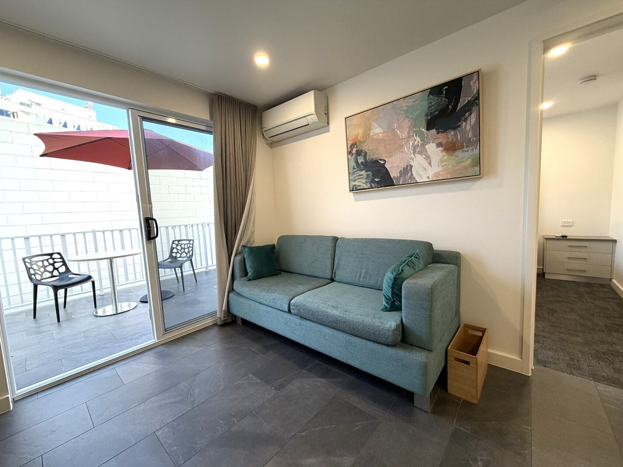 Una sala de estar moderna con un sofá gris, un ventanal que da a un área exterior con un paraguas rojo, y un cuadro en la pared.