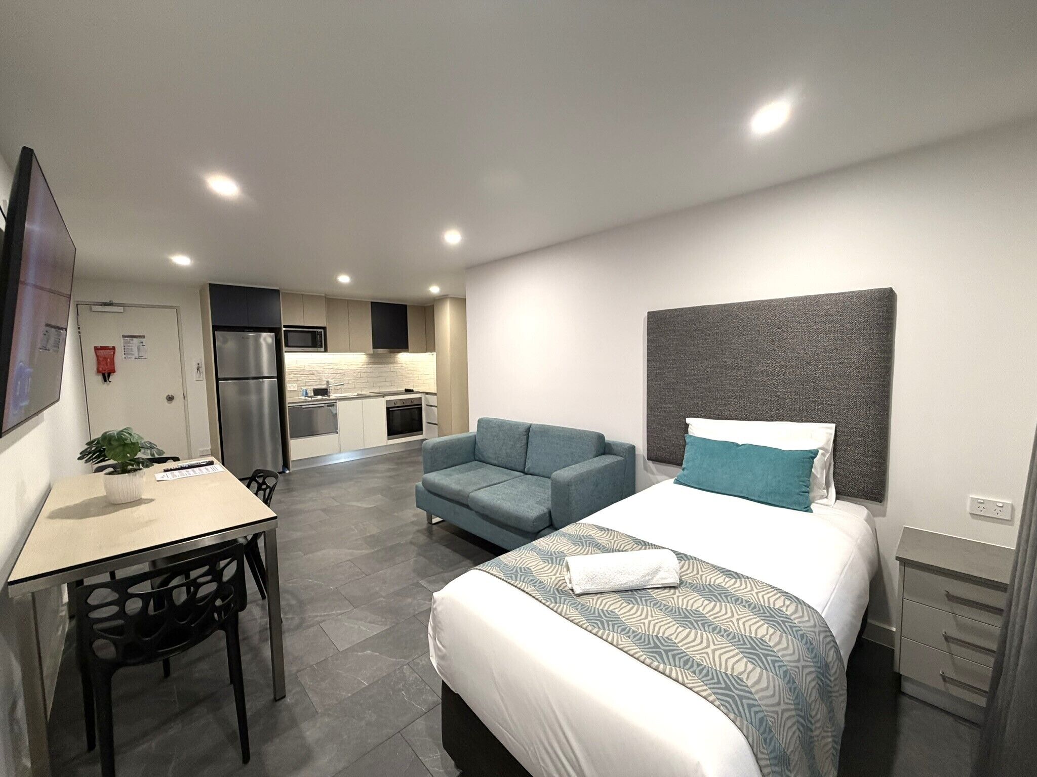Ein modernes Hotelzimmer mit Bett, Schreibtisch, Sessel und einer kleinen Küchenzeile im Hintergrund.
