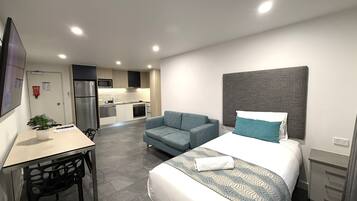 A modern hotel room with a bed, a desk, a chair, a sofa, and a kitchenette in the background.