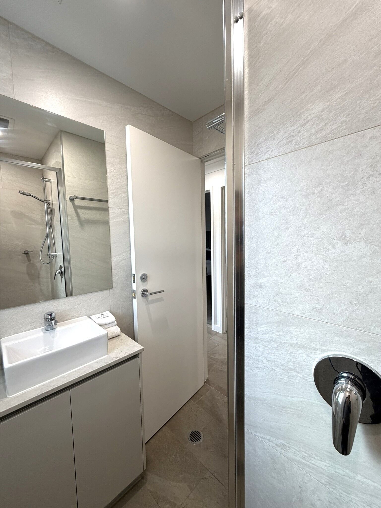 A modern bathroom with a white sink, a mirror, a shower, and a door leading to another room.