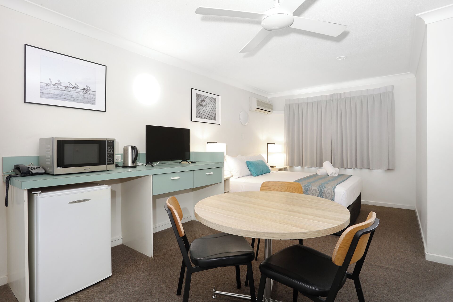 A compact hotel room with a kitchenette, a dining area, a bed, and a ceiling fan.