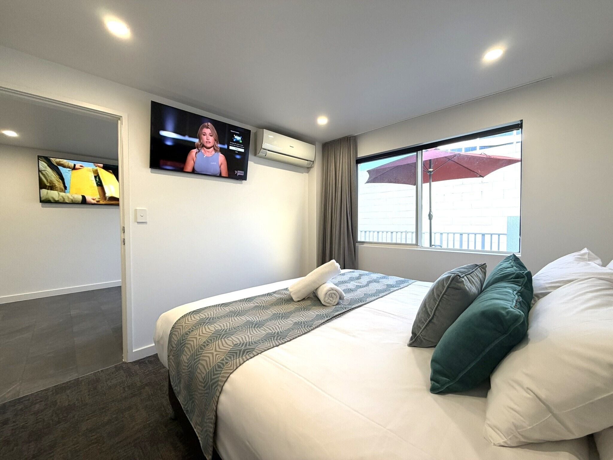 Una camera d'albergo con un letto, una TV fissata al muro, una finestra con un ombrello e una porta.