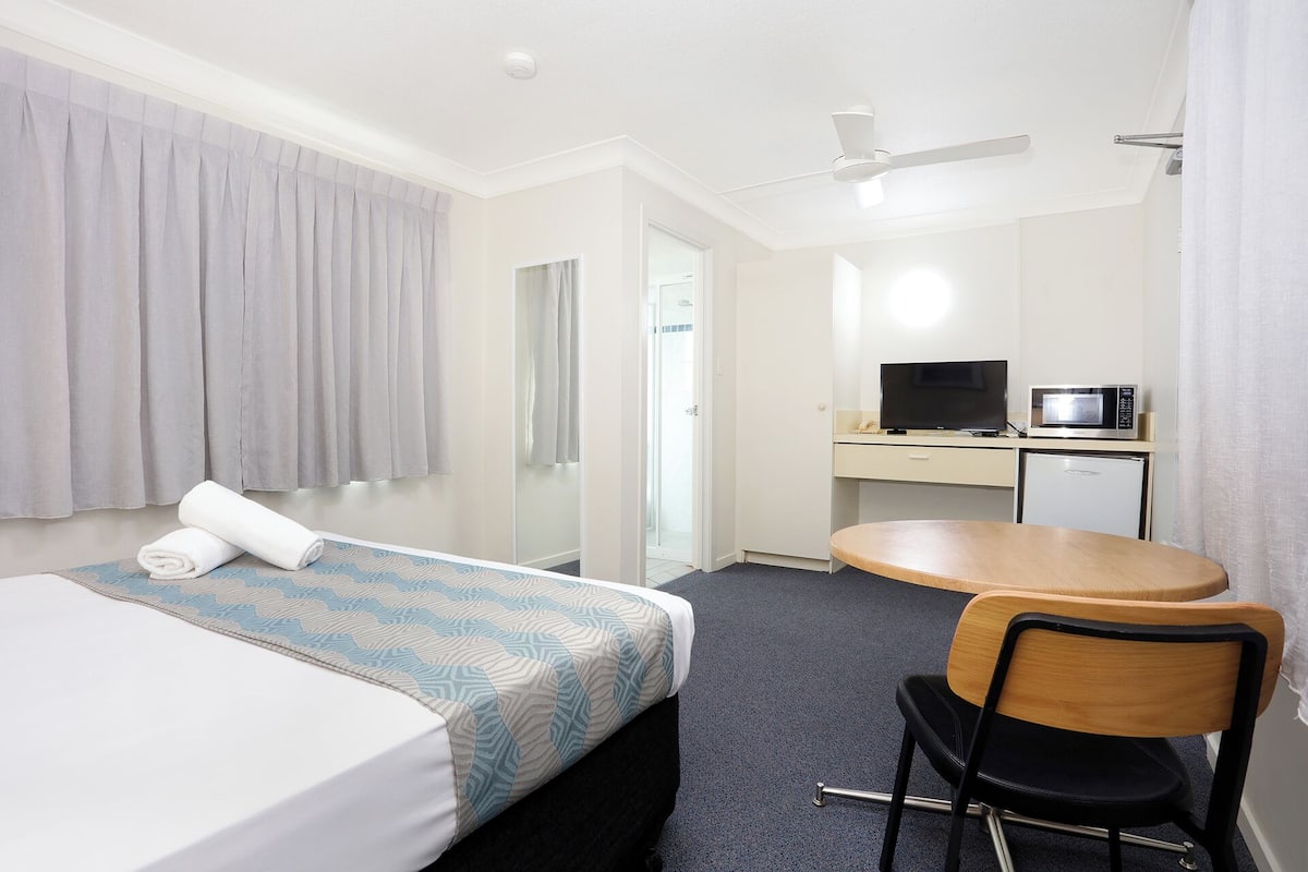 A hotel room with a bed, a desk with a television and microwave, a chair, and a ceiling fan.