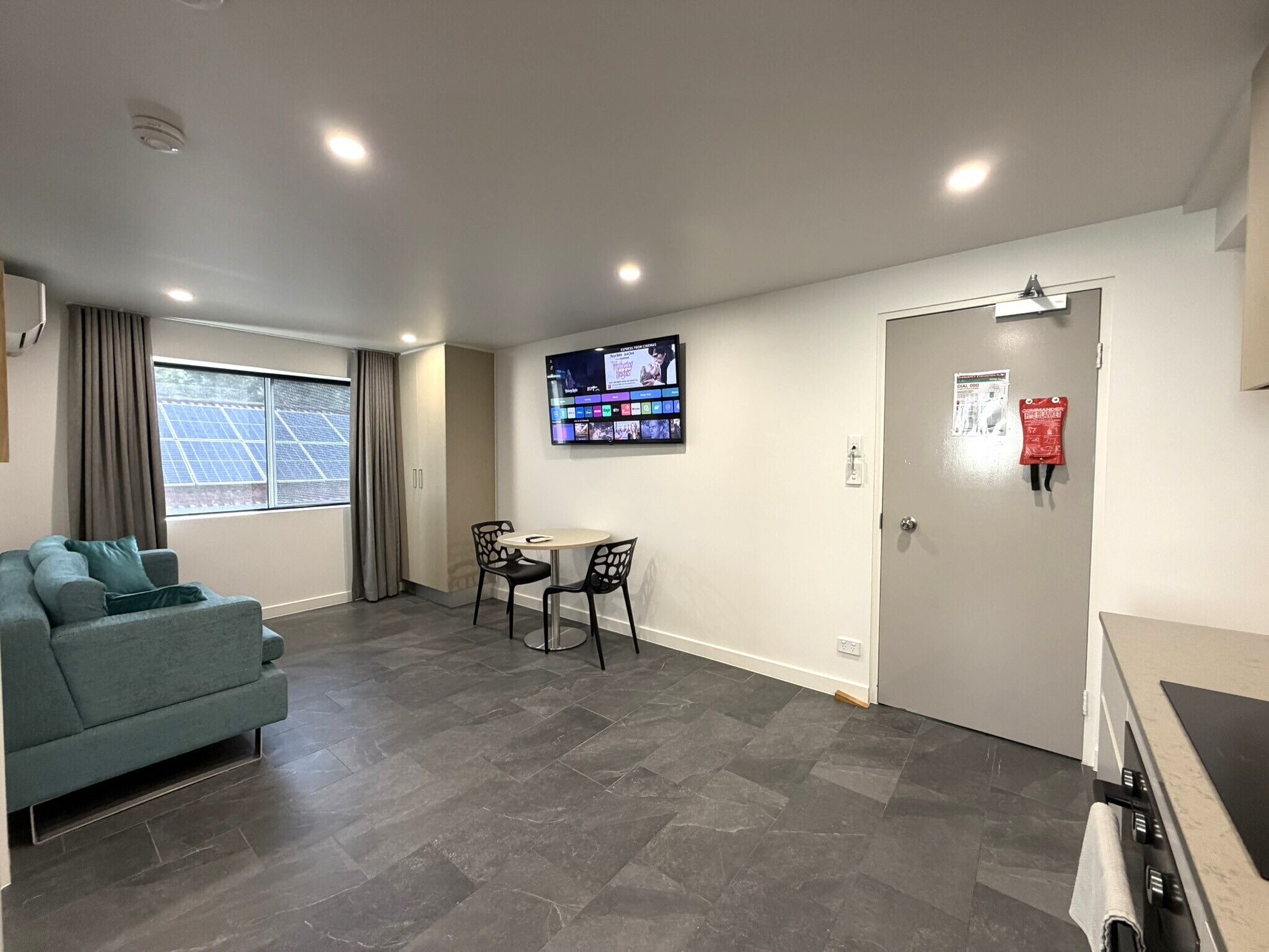 A modern hotel room with a sofa, a dining table with chairs, a TV mounted on the wall, and a door with a fire safety sign.
