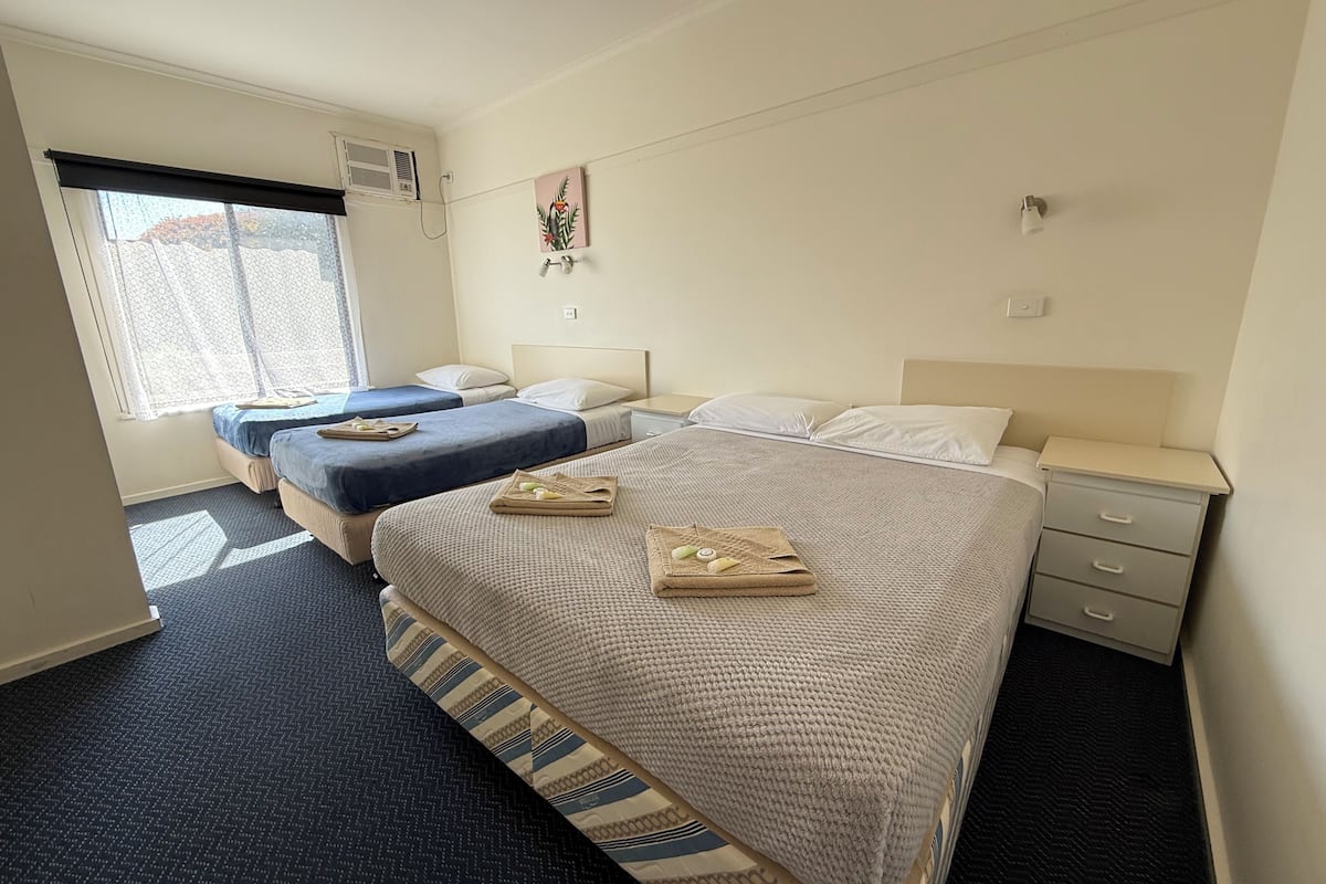 A hotel room with two single beds, a large bed, a window with a view, a painting on the wall, and a bedside table with a lamp.