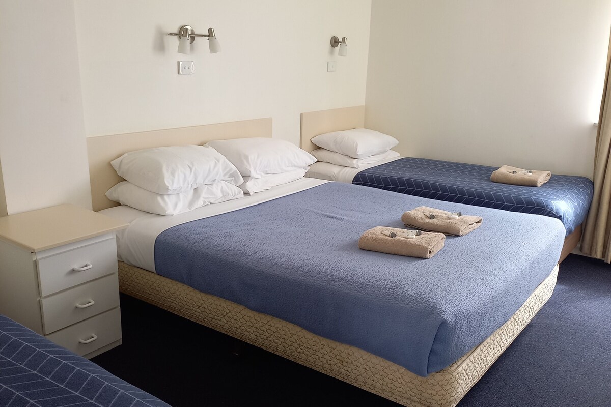 A hotel room with two beds, each with a blue bedspread and white pillows. There are two bedside tables with lamps, a wall-mounted air conditioner, and a window with curtains.