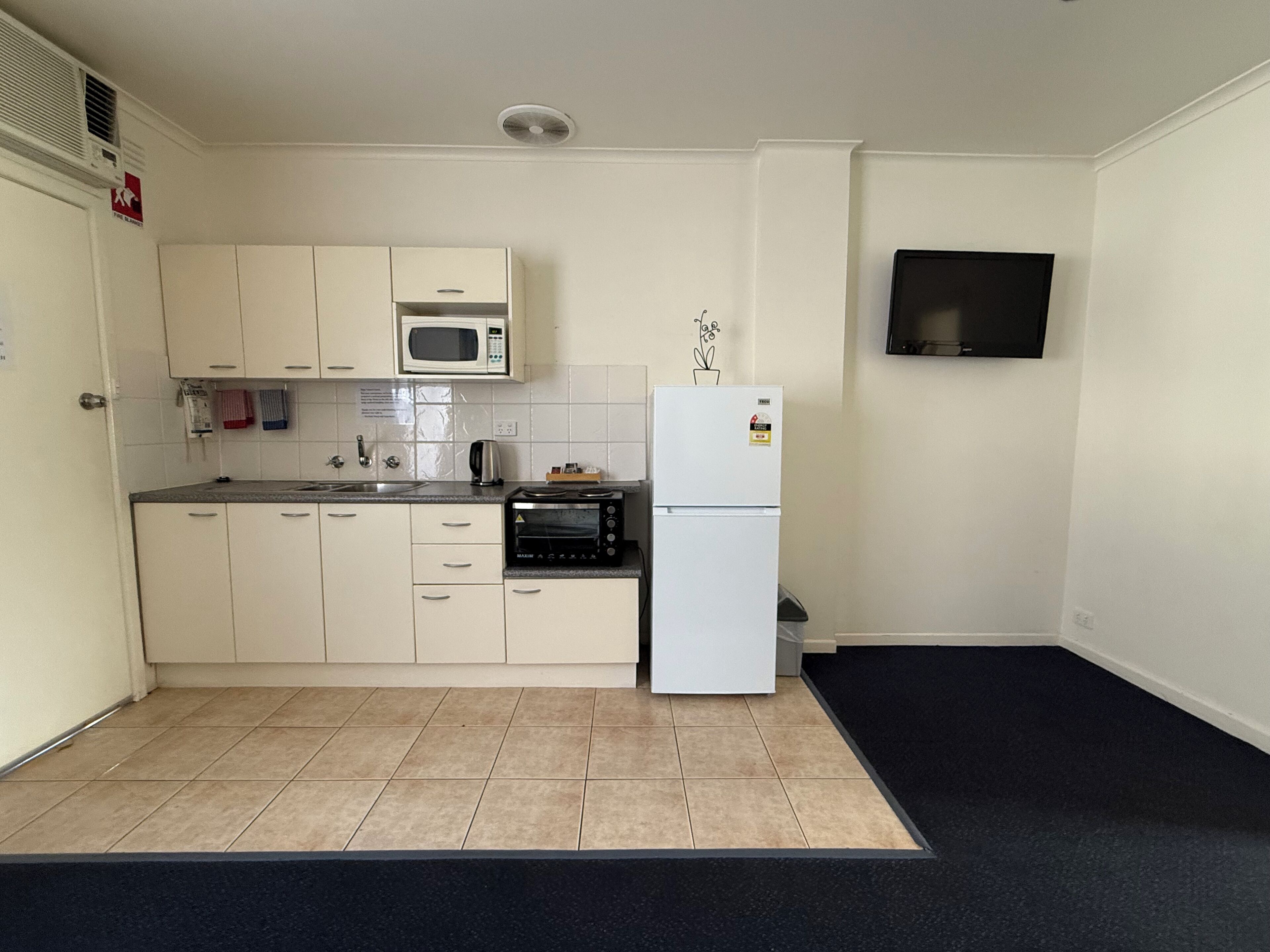 A compact kitchen with white cabinets, a microwave, a toaster, and a refrigerator. A flat-screen TV is mounted on the wall.