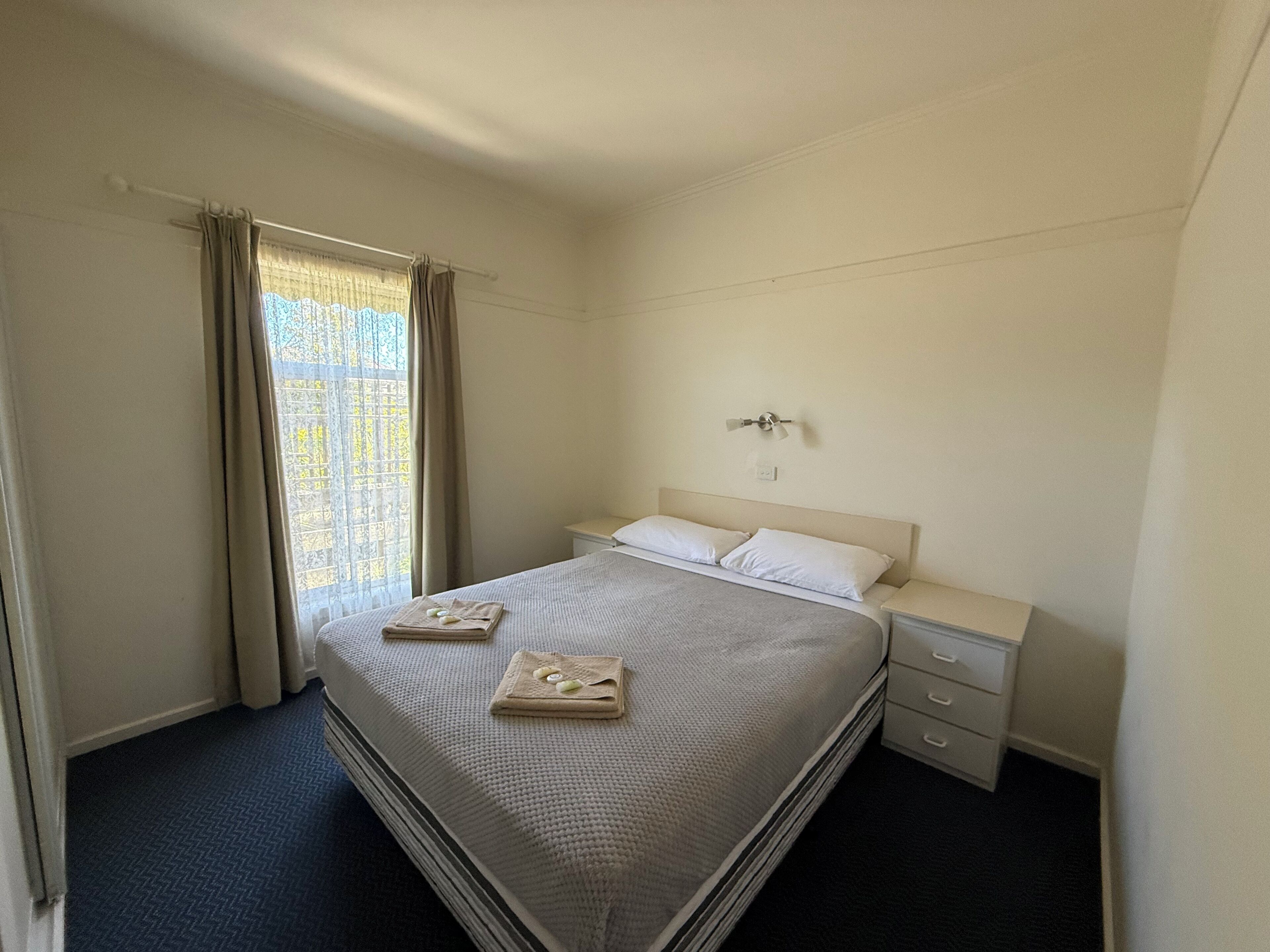 A hotel room with a bed, a nightstand, a wall-mounted fan, and a window with curtains.