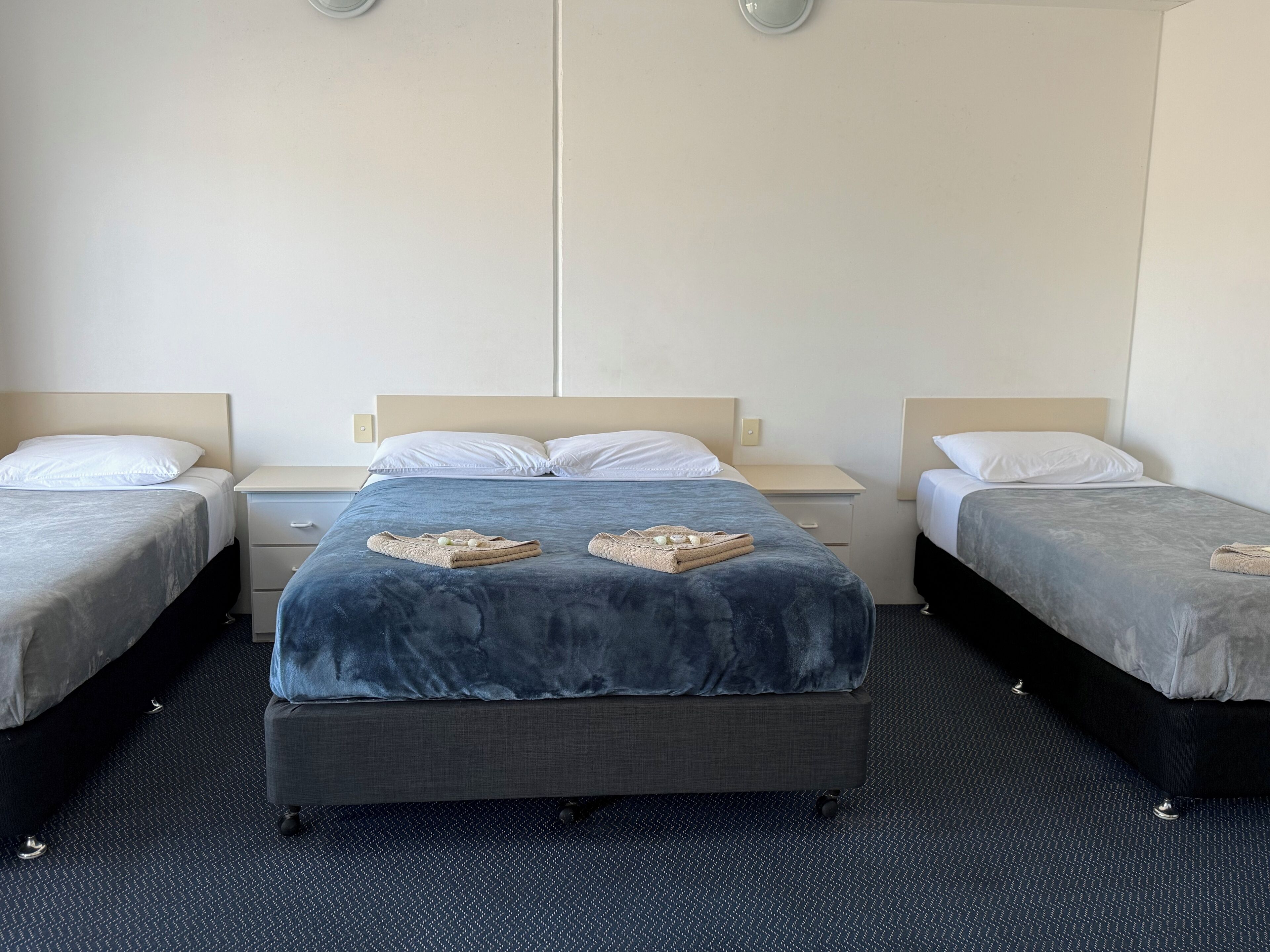 Habitación de hotel con tres camas individuales, cada una con cabecero gris y almohadas blancas. La cama central tiene una manta azul y dos cestas tejidas encima.