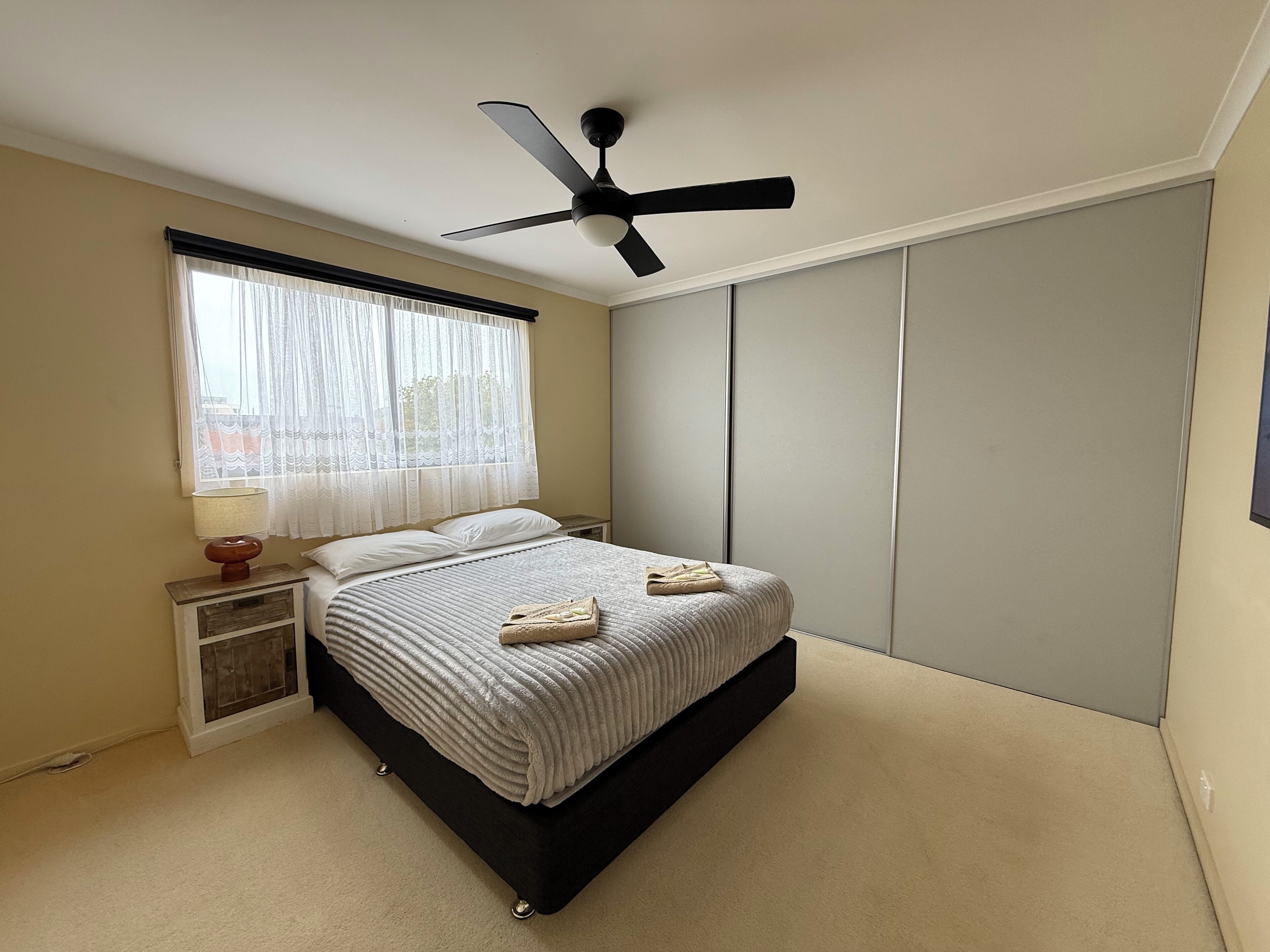 A bedroom with a bed, a ceiling fan, a nightstand with a lamp, a window with sheer curtains, and a wardrobe.