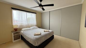 A bedroom with a bed, a ceiling fan, a nightstand with a lamp, a window with sheer curtains, and a wardrobe.