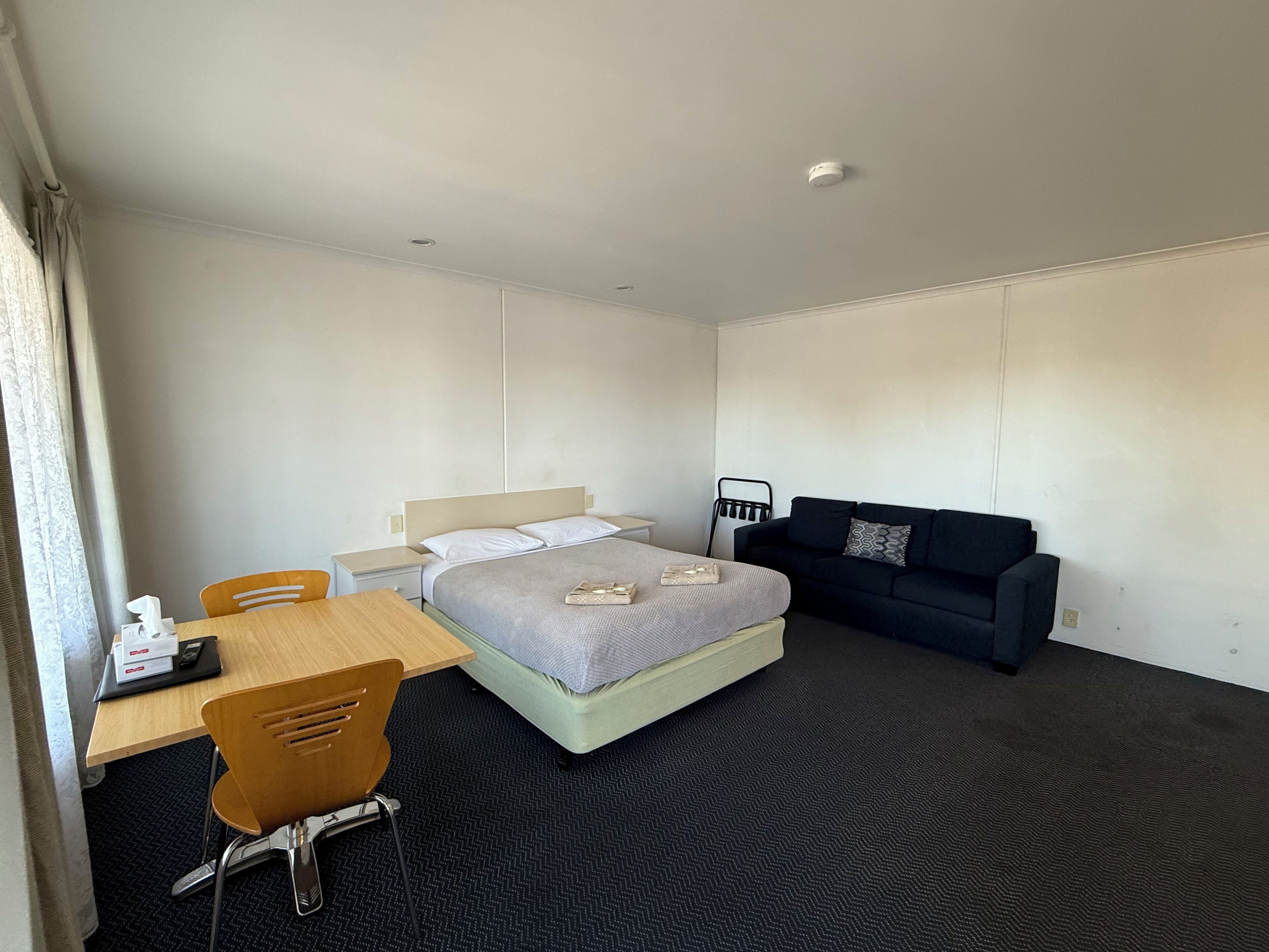 A hotel room with a bed, a desk with a chair, a small table, a sofa, and a window with curtains.