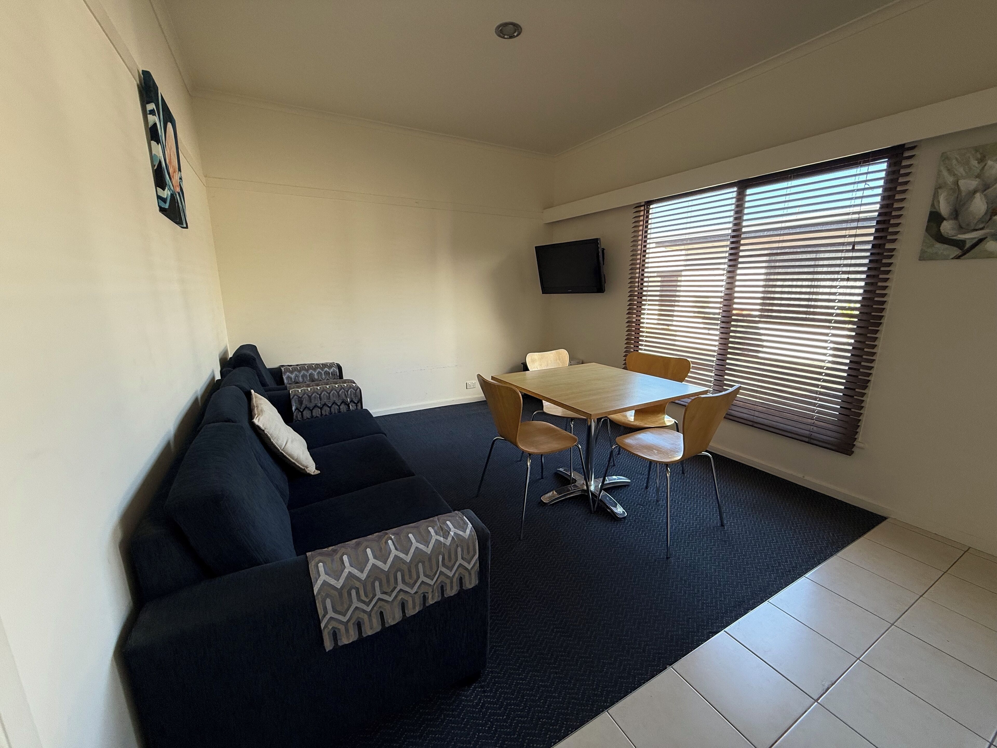 A living room with a dark blue sofa, a wooden dining table with four chairs, a flat-screen TV mounted on the wall, and a window with blinds.