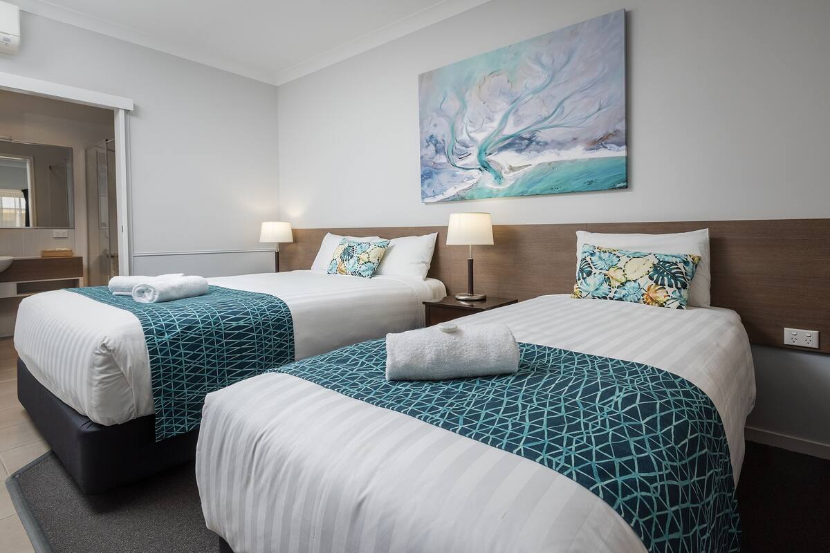 A hotel room with two beds, each with a geometric patterned bedspread, a large abstract painting on the wall, and a bathroom visible through an open door.