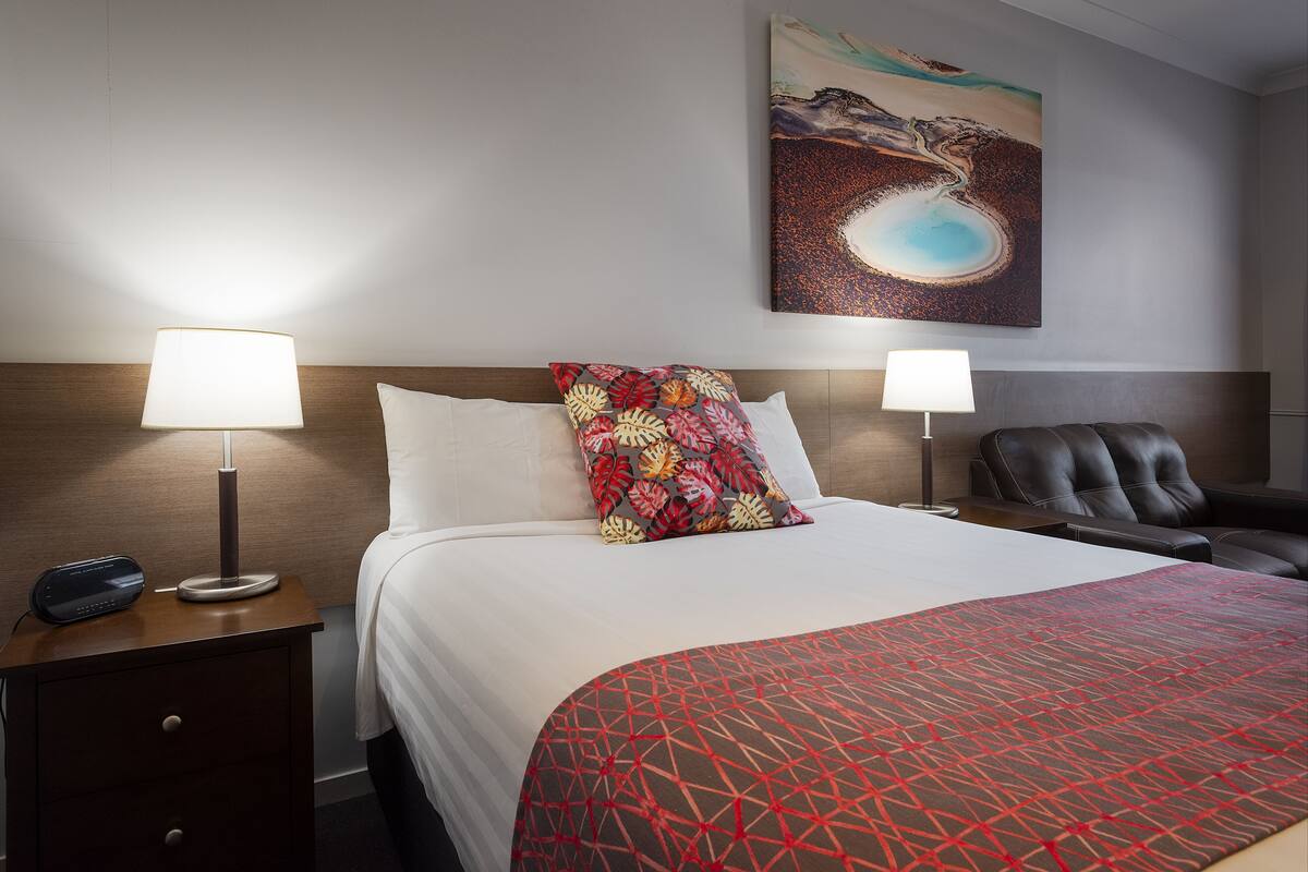 A hotel room with a bed, two bedside lamps, a chair, a nightstand, and a painting on the wall.