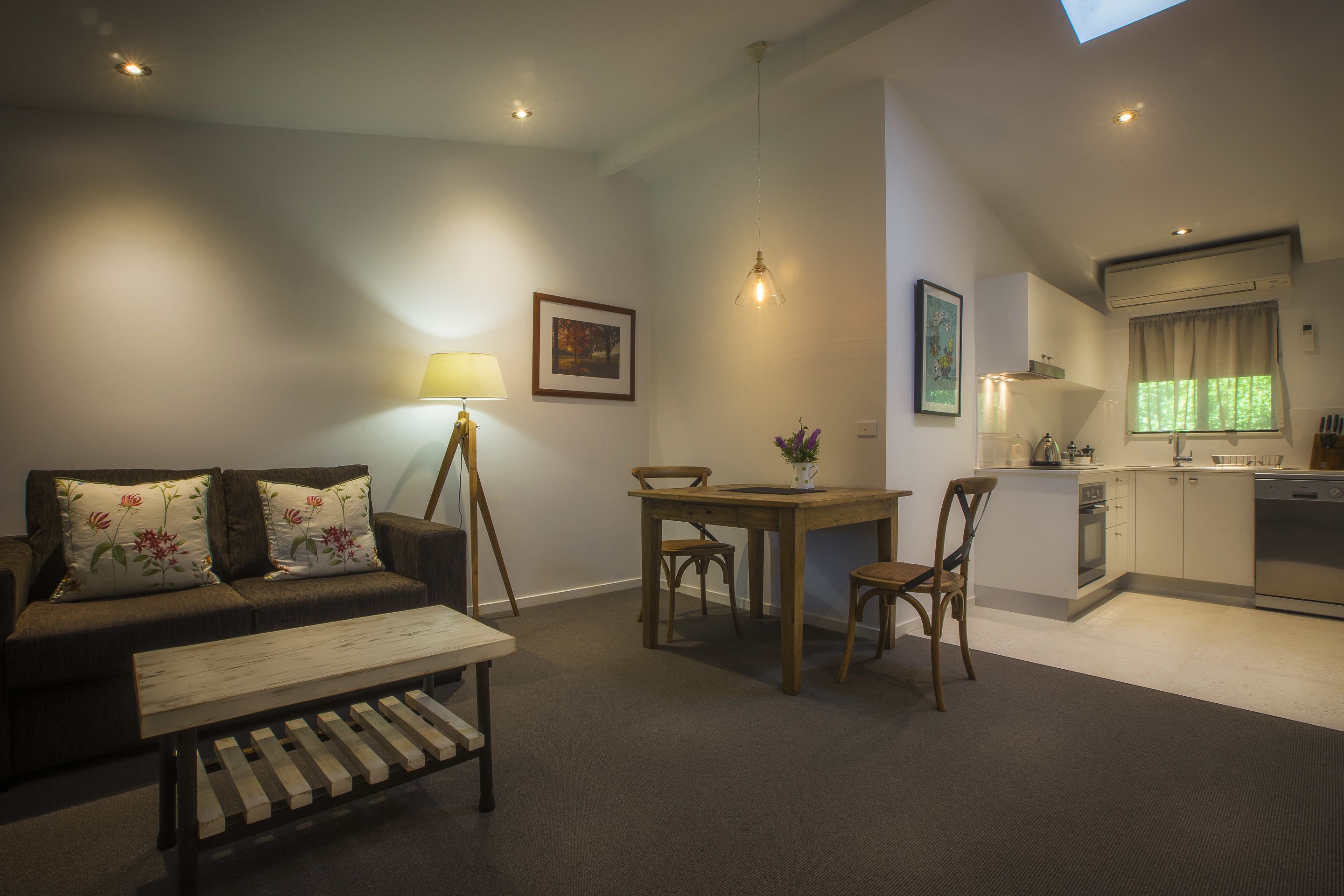 A compact living space with a sofa, a wooden coffee table, a dining table with chairs, a kitchen area with cabinets and appliances, and a skylight.