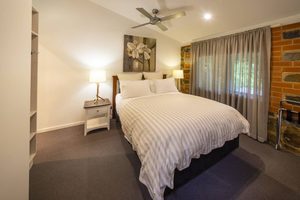 A bedroom with a bed, nightstands, a ceiling fan, a painting, and a window with curtains.
