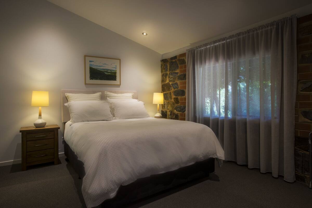 A bedroom with a large bed, two bedside tables with lamps, a stone accent wall, and a window with curtains.