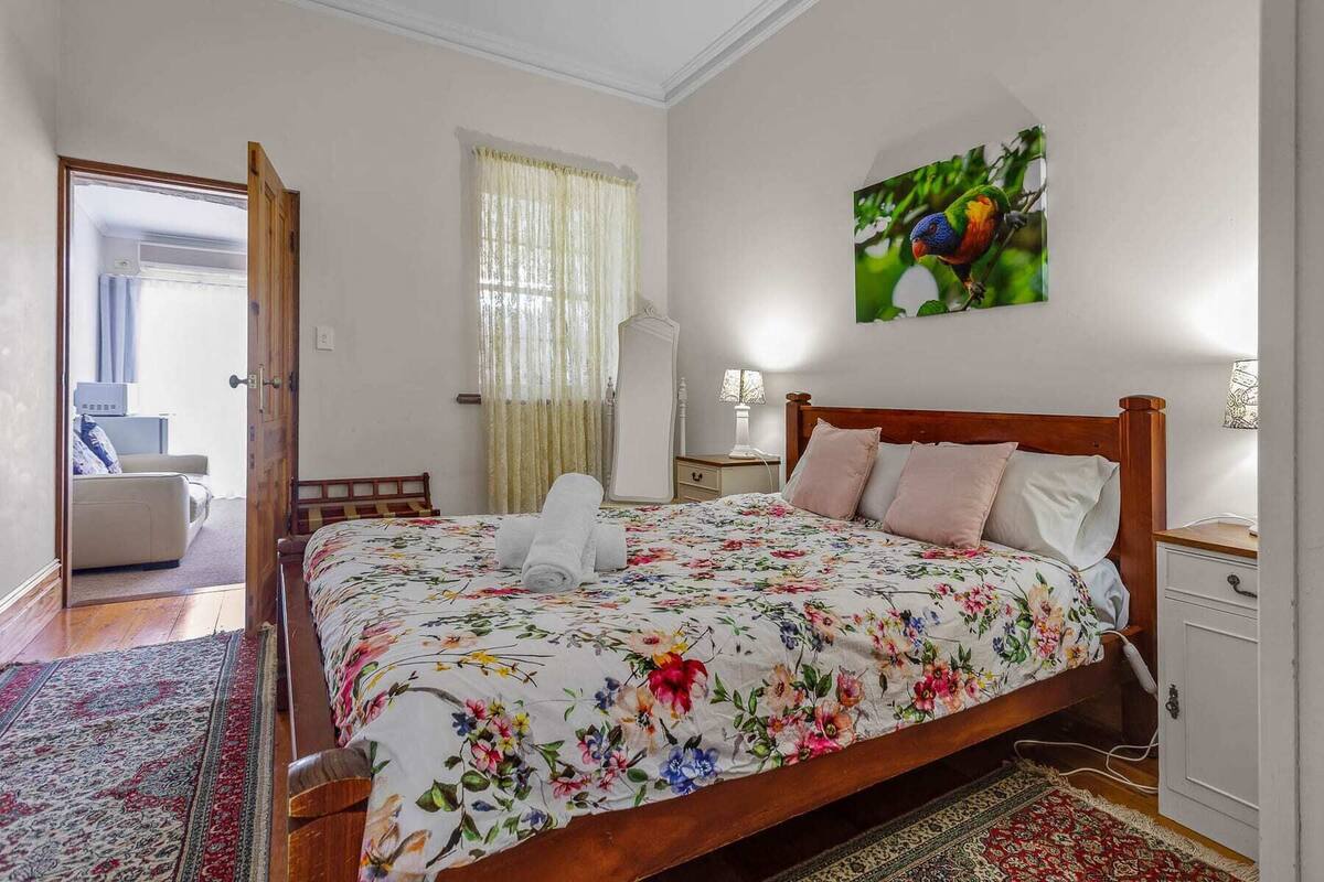 Une chambre à coucher avec un lit en bois, une couvre-lit à motifs floraux, une table de chevet avec une lampe, une commode, une fenêtre avec des rideaux légers et un tableau représentant un oiseau.