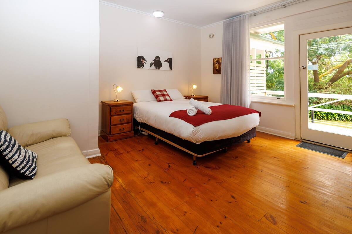 Une chambre avec un plancher en bois, un lit recouvert d’une couverture rouge, une table de chevet, une lampe, une chaise et une fenêtre donnant sur un paysage verdoyant.