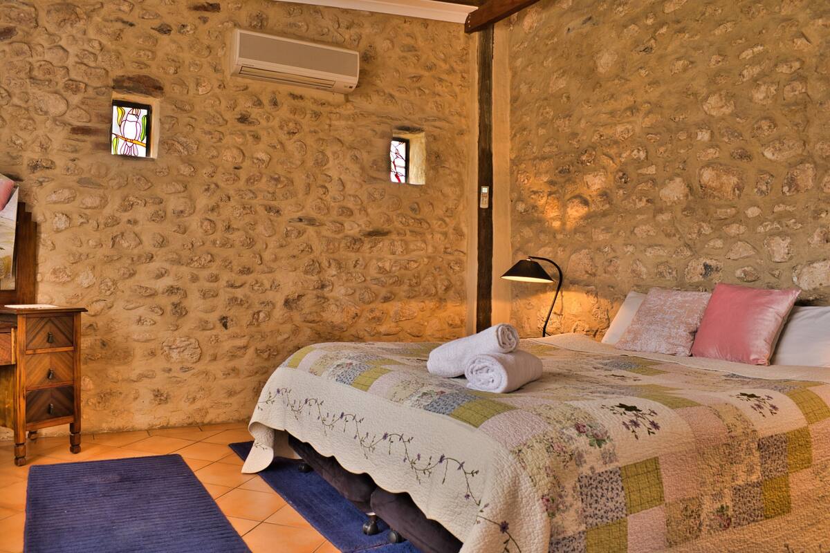 Une chambre aux murs de pierre, avec une couvre-lit matelassé, une table de chevet en bois et un tapis bleu.