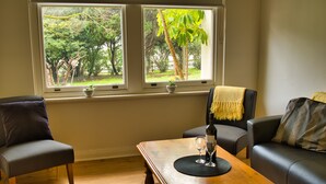 Un salon comprenant deux fauteuils, une table basse en bois et un canapé. Il y a deux fenêtres donnant sur un paysage verdoyant.