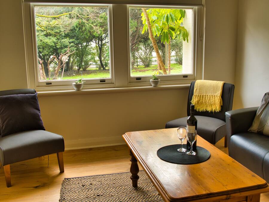 A living room with two armchairs, a wooden coffee table, and a sofa. There are two windows with a view of greenery outside.